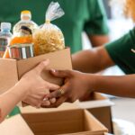 Volunteers wearing green shirts hand over a box of food and bottled water, representing kindness and teamwork in charity work.