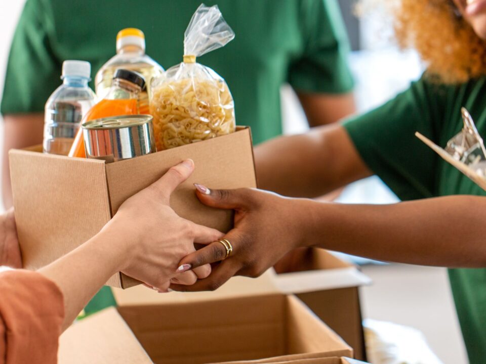 Volunteers wearing green shirts hand over a box of food and bottled water, representing kindness and teamwork in charity work.