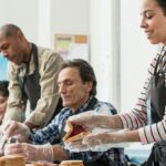 A diverse team of four adults works together to prepare food. The woman in the foreground is carefully making a jam and peanut butter sandwich while wearing an apron and clear plastic gloves, demonstrating safe food handling for a nonprofit organization's charity meal program.