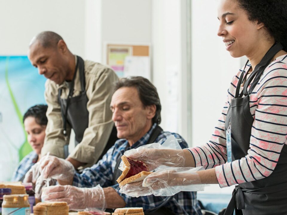 A diverse team of four adults works together to prepare food. The woman in the foreground is carefully making a jam and peanut butter sandwich while wearing an apron and clear plastic gloves, demonstrating safe food handling for a nonprofit organization's charity meal program.